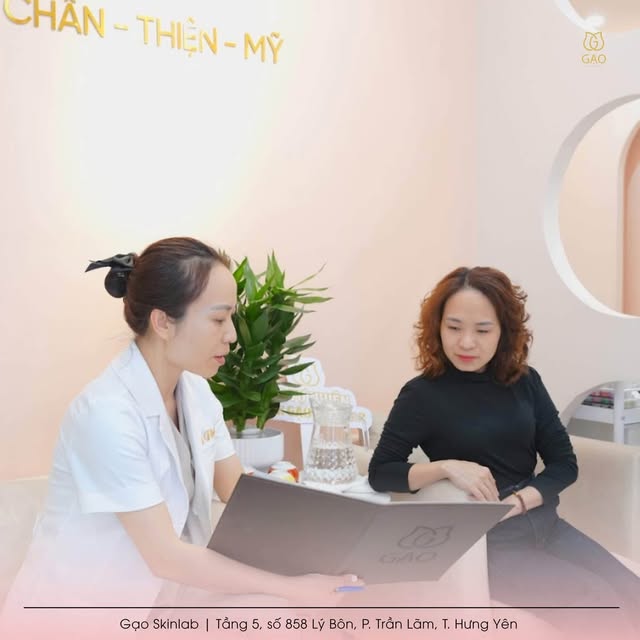 Ảnh từ fanpage Gạo Skinlab: người tư vấn áo trắng cầm cuốn sổ Gạo trò chuyện cùng khách trên sofa, phía sau là dòng chữ Chân – Thiện – Mỹ trên tường.